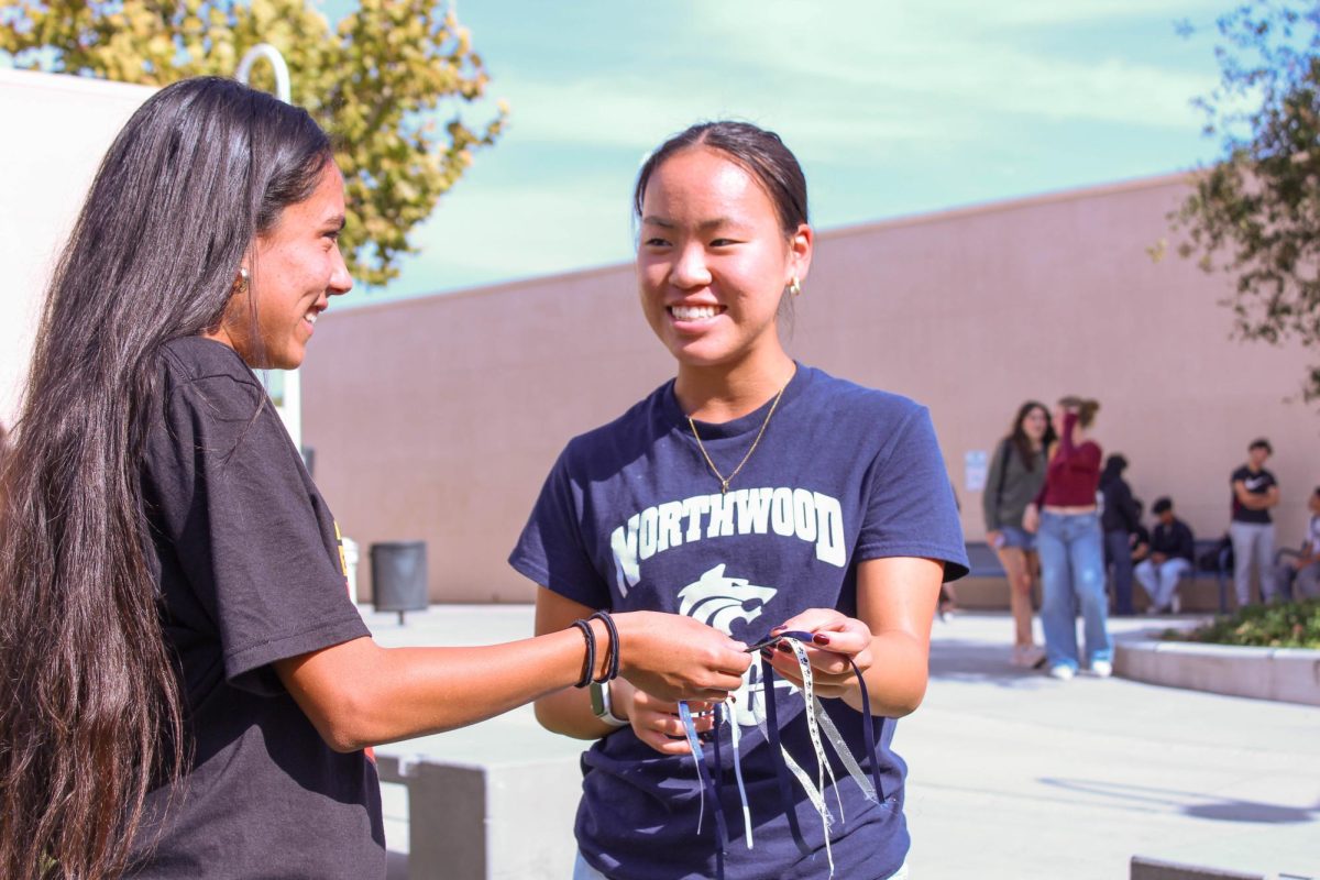 SPRINTING WITH STYLE: Cross country captain senior Christine Nam delivers her handmade and custom blue and silver hair ribbons to teammate sophomore Braelynn Lynch in preparation for an upcoming cross country championship meet.